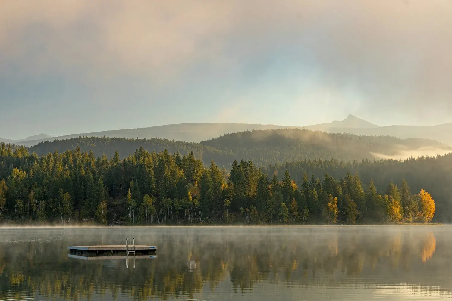A still lake at dawn, mist rising off the water, a small dock at the edge, forested hills beyond.